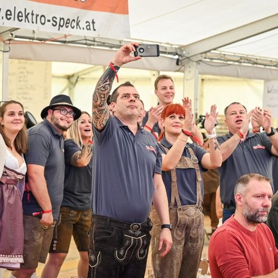 Eine Gruppe von Menschen in traditioneller bayerischer Kleidung klatscht und jubelt in einem Zelt im Freien. Ein Mann in der Mitte macht ein Selfie mit einer Kamera.