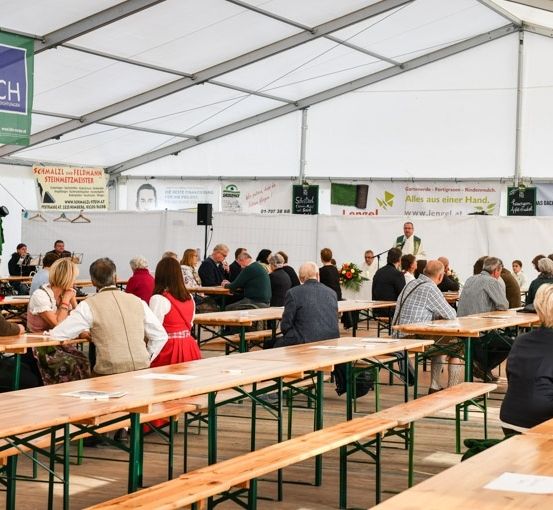 Eine große Gruppe von Menschen sitzt an langen Holztischen in einem Zelt. Ein Mann steht vorne und spricht die Zuhörer an. Banner und Poster sind an den Zeltwänden angebracht.