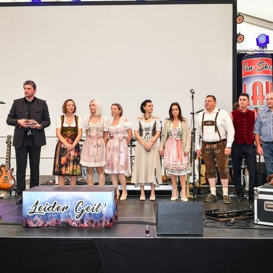 Eine Gruppe von Menschen in traditioneller Kleidung steht auf der Bühne mit einem Mann, der ein Mikrofon hält. Hinter ihnen steht eine große Leinwand, ein Lautsprecher und eine Gitarre. Die Bühne hat ein Schild mit der Aufschrift Leider Geil!