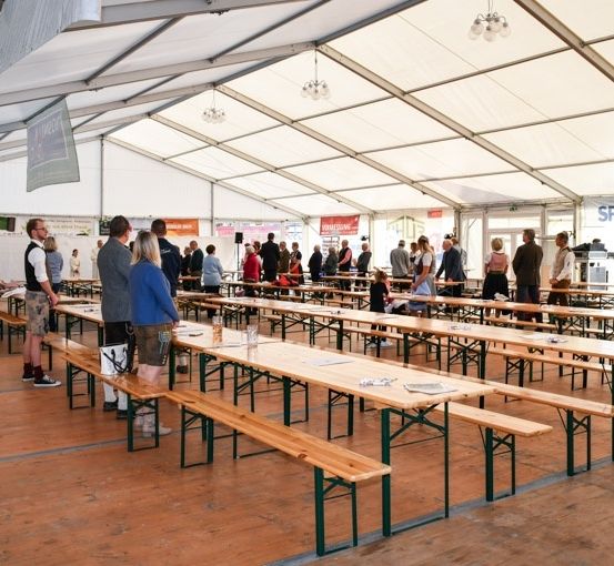 Ein großes Zelt mit hölzernen Bänken und Tischen für eine Veranstaltung, mit vielen Menschen, die stehen und sich unterhalten. Das Zelt hat weiße Wände und ein Metallgerüst, mit Lampen, die von der Decke hängen.