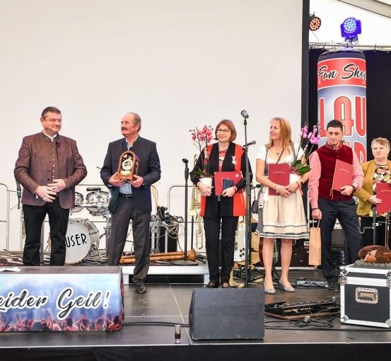 Eine Gruppe von Menschen steht auf einer Bühne, hält Auszeichnungen und Blumen. Ein Mann mit einem Mikrofon steht links, und ein Schlagzeug steht hinter ihnen.