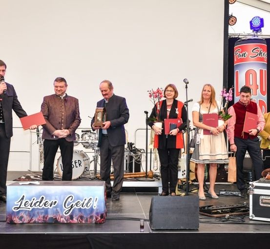 Eine Gruppe von Menschen auf der Bühne, einige halten rote Ordner, ein Mann hält ein Mikrofon, und ein Schild, das Leider Geil! steht. Ein Schlagzeug und Gitarren sind hinter ihnen.