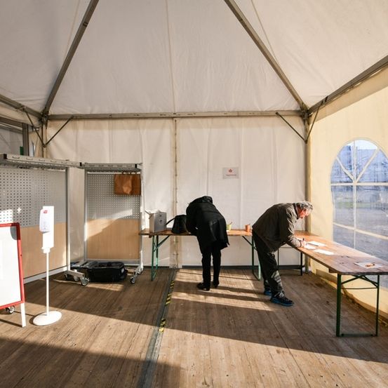 Zwei Personen stehen in einem Zeltwahlbereich, eine schreibt an einem Schreibtisch, während die andere in der Nähe steht. Es gibt eine Tafel, einen Schreibtisch und eine Tasche auf dem Boden.