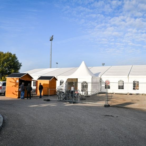 Ein großes weißes Zelt steht in einem offenen Bereich mit einem Metallzaun. Zwei Personen stehen in der Nähe einer kleinen orangefarbenen Struktur. Fahrräder sind vor dem Zelt geparkt. Bäume und ein weißer Van sind im Hintergrund sichtbar.