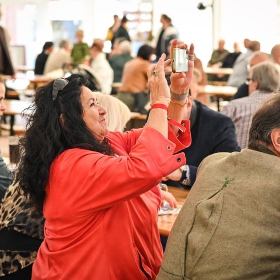 Eine Frau in einem roten Mantel macht ein Foto mit ihrem Handy bei einer überfüllten Indoor-Veranstaltung.