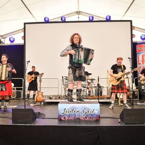 Eine Gruppe Musiker tritt auf der Bühne auf, wobei ein Mitglied ein Akkordeon spielt, während andere Gitarren und Schlagzeug spielen. Die Bühne ist mit Lautsprechern und einer großen Leinwand im Hintergrund ausgestattet.