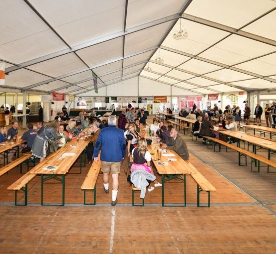 Ein großer Innenbereich mit langen Holztischen und Bänken, gefüllt mit sitzenden und stehenden Personen. Der Raum befindet sich unter einem weißen Zelt mit Bannern und Schildern.