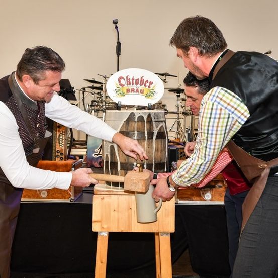 Drei Männer in traditioneller Kleidung klopfen mit einem Hammer auf einen Fass, einer hält einen Becher. Im Hintergrund sind Musikinstrumente zu sehen.