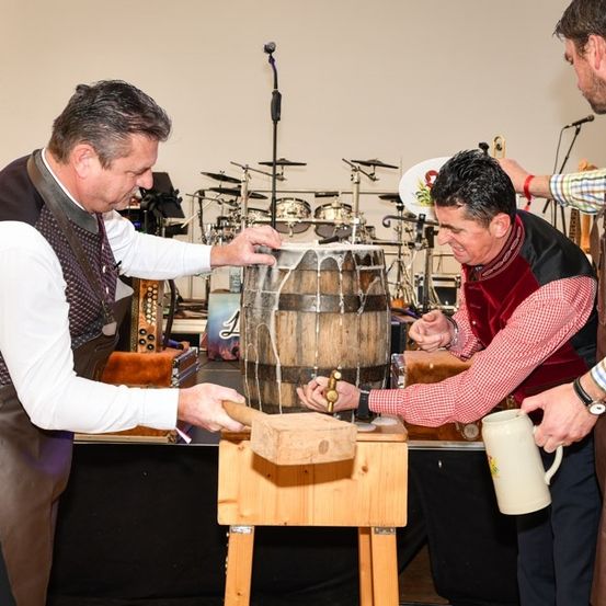 Drei Männer klopfen mit einem Hammer auf einen Holzfass auf der Bühne, einer hält einen Krug. Im Hintergrund steht ein Schlagzeug und Mikrofone.