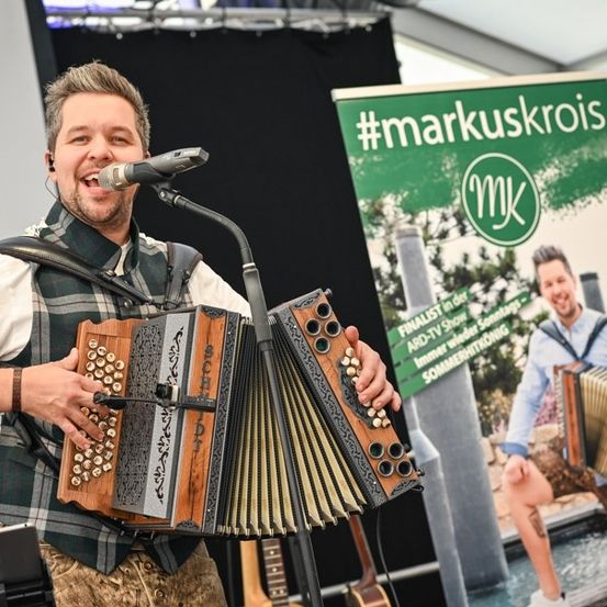Ein Mann in einem karierten Hemd und einer Hose mit Hosenträgern spielt Akkordeon und singt in ein Mikrofon bei einem Musikevent. Hinter ihm ist ein Banner mit dem Hashtag #markuskrois und einem Mann, der Akkordeon spielt, zu sehen.
