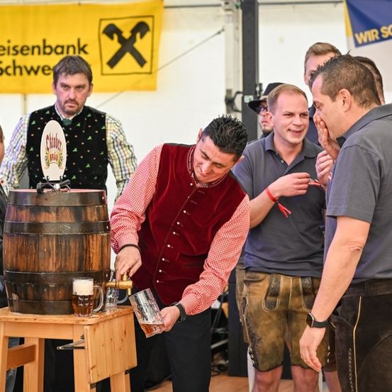Ein Mann zapft ein Bier aus einem Holzfass in ein Glas auf einem Holztisch, während andere zusehen.