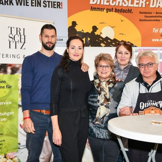 Fünf Personen stehen zusammen vor einem Banner mit einer Weinflasche auf einem Tisch. Das Banner hat den Text 'Drechler Dach' und 'Trischitz Catering'. Die Leute lächeln und scheinen für ein Foto zu posieren.