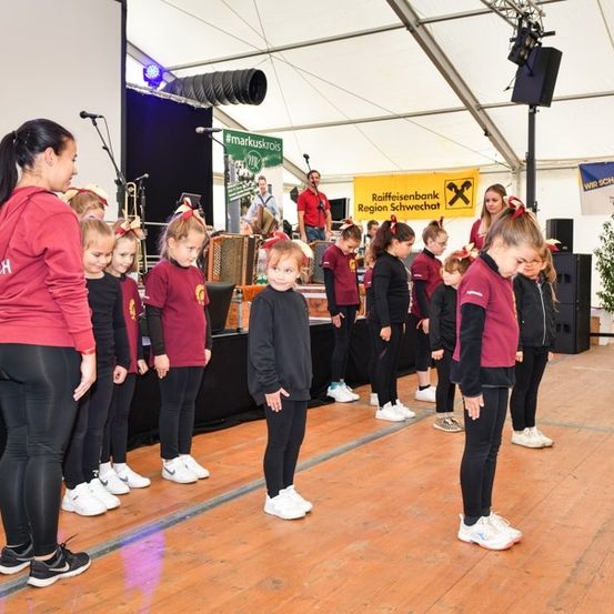 Eine Gruppe junger Mädchen in roten und schwarzen Uniformen steht auf der Bühne mit einer Frau vor ihnen. Dahinter spielt eine Band auf der Bühne mit einem gelben Banner und einem Topf.