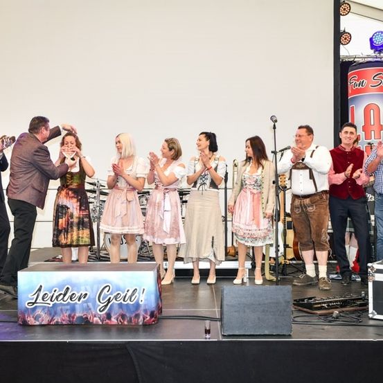 Eine Gruppe von Musikern und Darstellern auf der Bühne, einige klatschen und andere halten Instrumente, mit einem Banner, das Leider Geil! vor ihnen steht.
