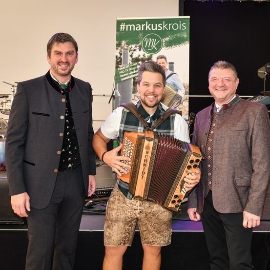 Drei Männer stehen auf einer Bühne, einer hält ein Akkordeon. Hinter ihnen ist ein Banner mit dem Hashtag #markuskrois. Der Mann links trägt einen Anzug, der Mann in der Mitte ein traditionelles Outfit und der Mann rechts einen braunen Mantel. Im Hintergrund sind Musikinstrumente zu sehen.