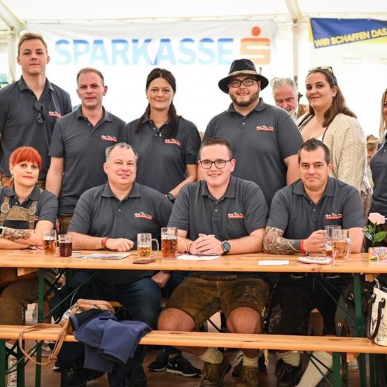 Eine Gruppe von Menschen in passenden Shirts sitzt an einem Tisch mit Bierkrügen und lächelt für ein Foto. Der Banner hinter ihnen lautet 'SPARKASSE'.