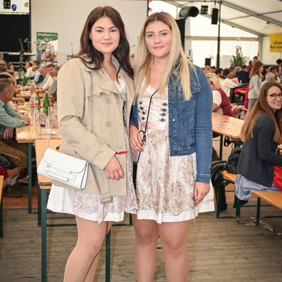 Zwei Frauen stehen in einem Zelt-Event im Freien, eine mit einer beigefarbenen Trenchcoat und die andere in einer Jeansjacke. Sie sind von Tischen und Menschen umgeben.