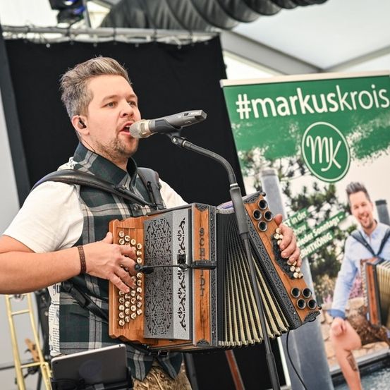 Ein Mann spielt Akkordeon und singt in ein Mikrofon auf der Bühne, mit einem Banner für #markuskrois im Hintergrund.