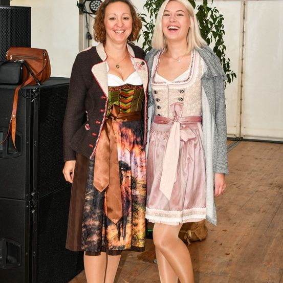 Zwei Frauen stehen lächelnd vor einem Hintergrund mit einem Banner und Lautsprechern. Eine trägt ein traditionelles deutsches Kleid, die andere ein lässiges Outfit.