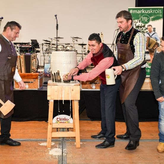 Vier Männer auf einer Bühne, zwei halten einen Hammer und einen Becher, die anderen zwei halten einen großen Fass, möglicherweise an einer traditionellen Veranstaltung teilnehmend.