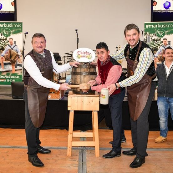 Fünf Männer in traditioneller Kleidung stehen auf der Bühne, zwei von ihnen klopfen mit einem Hammer auf einen Holzfass. Der Mann in der Mitte hält einen Krug. Hinter ihnen ist ein Banner mit dem Hashtag #markuskrois zu sehen.