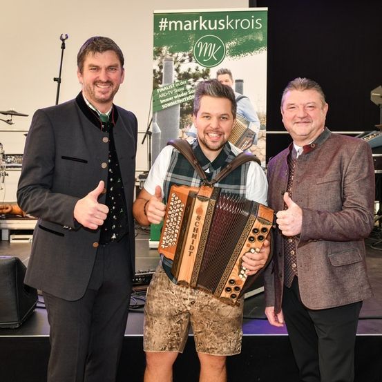 Drei Männer auf der Bühne, einer hält ein Akkordeon, lächeln und geben Daumen hoch. Hinter ihnen ist ein Banner mit dem Text '#markuskrois'.