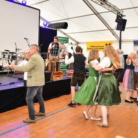 Eine Gruppe von Menschen tanzt auf einer Bühne mit einer Band in einem Zelt, ein Mann spielt Akkordeon, während andere in traditioneller Kleidung tanzen.