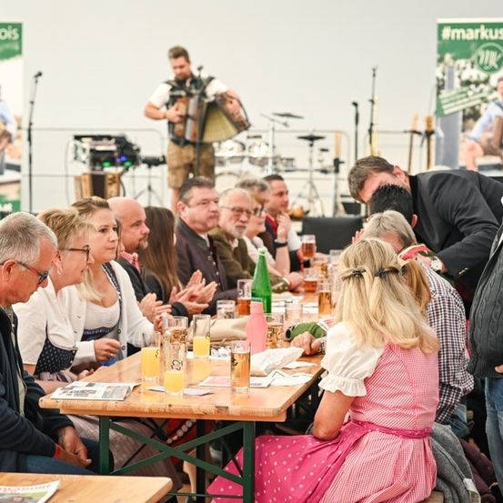 Eine Gruppe von Menschen sitzt an einem Tisch mit Getränken und Essen, während ein Mann auf der Bühne ein Akkordeon spielt. Ein Banner mit dem Text #markuskrois ist im Hintergrund zu sehen.
