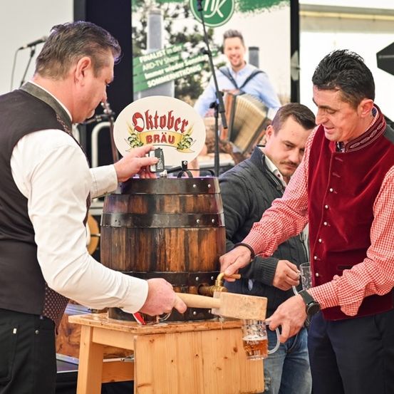 Zwei Männer klopfen mit einem Hammer auf einen Holzfass, um Bier in ein Glas zu gießen, mit einem Schild im Hintergrund, das 'Oktoberbrau' steht.