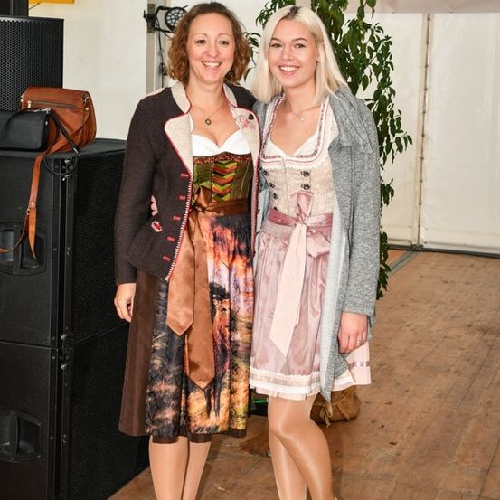 Zwei Frauen in traditioneller deutscher Kleidung, die nebeneinander stehen und lächeln, mit einem großen Lautsprecher und einer Pflanze im Hintergrund.