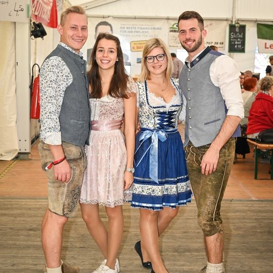 Drei junge Erwachsene, gekleidet in traditioneller deutscher Kleidung, stehen in einer festlichen Umgebung zusammen und lächeln für die Kamera. Der Mann links trägt eine Weste und Ledershorts, die Frau in der Mitte trägt ein weißes Kleid und der Mann rechts hat eine Weste und Hose an. Hinter ihnen befinden sich Banner und ein verschwommener Hintergrund von Menschen.