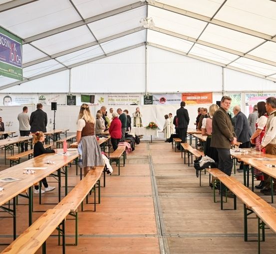 Ein großer Innenbereich mit Holzbänken und Tischen, wo sich Menschen versammeln. Einige stehen, andere sitzen, und es hängen Banner von der Decke.