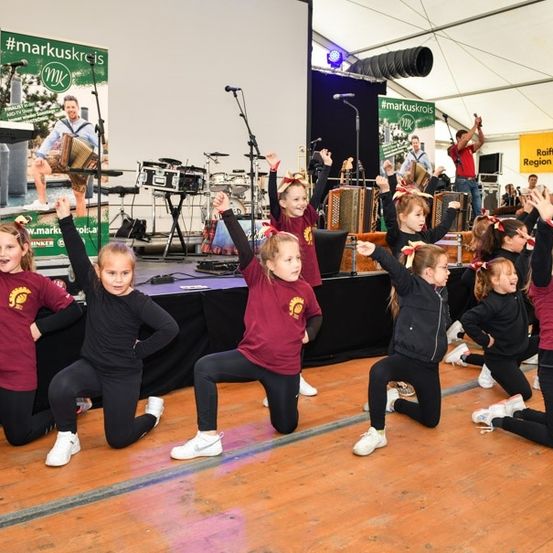 Eine Gruppe junger Mädchen in roten und schwarzen Outfits tritt auf der Bühne auf, mit Musikinstrumenten im Hintergrund.