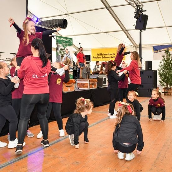 Eine Gruppe junger Mädchen in passenden roten und schwarzen Outfits führt eine Cheerleading-Routine auf der Bühne auf. Dahinter spielt ein Mann Schlagzeug.