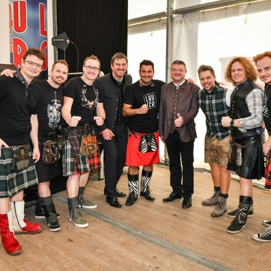 Eine Gruppe von Männern, die in Kilts und verschiedenen Accessoires gekleidet sind, posiert für ein Foto auf einer Bühne. Sie lächeln und geben Daumen hoch.