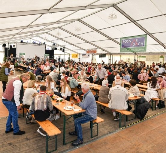 Eine große Gruppe von Menschen isst in einem Zeltbereich, mit Tischen und Bänken für die Sitzplätze. Einige stehen und unterhalten sich, während andere sitzen und essen. Das Zelt hat Banner und Lichter.