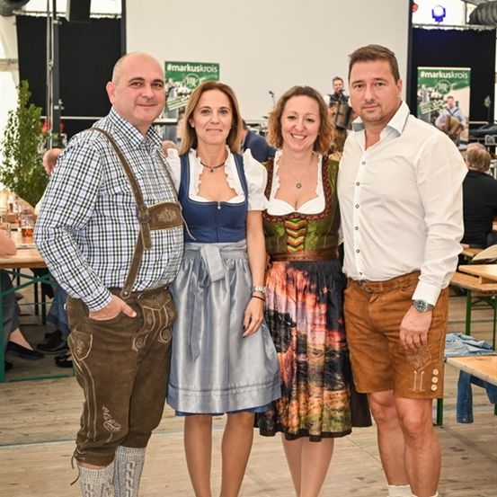 Drei Personen in traditioneller bayerischer Kleidung, die für ein Foto in einer festlichen Umgebung posieren, mit einer Bühne und Publikum im Hintergrund.