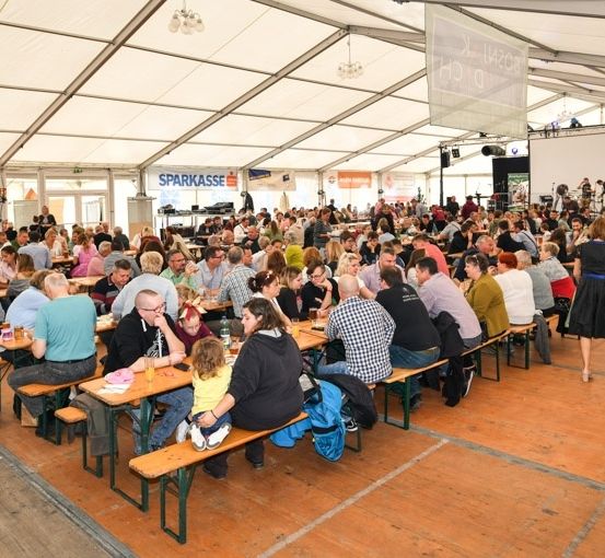 Eine große Gruppe von Menschen versammelt sich in einem Zelt zum Essen, viele sitzen an Holztischen und Bänken. Das Zelt ist gut beleuchtet, und im Hintergrund sind Lautsprecher und eine Bühne zu sehen.