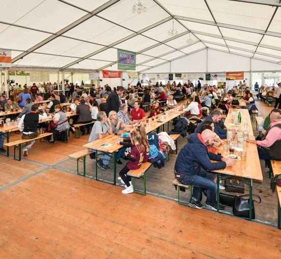 Eine große Gruppe von Menschen sitzt an Tischen in einem Zeltbereich, möglicherweise bei einem Festival. Die Tische sind mit verschiedenen Gegenständen gefüllt, einschließlich Flaschen und Gläser. Das Zelt wird von Metallrahmen gestützt, und es hängen Banner und Schilder von der Decke.
