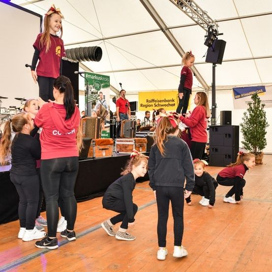 Eine Gruppe junger Mädchen in passenden roten und schwarzen Outfits tritt auf einer Bühne mit Musikinstrumenten und einem Banner im Hintergrund auf.