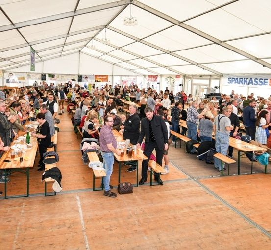 Eine große Menschenmenge versammelt sich in einem Zelt, sitzt an Tischen und Bänken. Einige essen und trinken, während andere stehen und reden. Das Zelt hat eine weiße Decke und Wände mit Bannern.