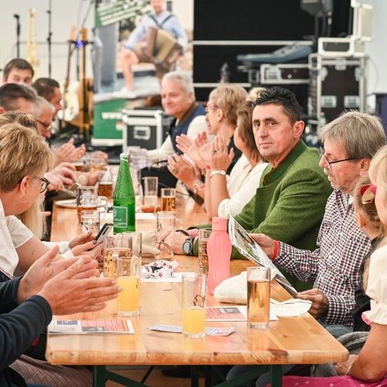 Eine Gruppe von Menschen sitzt um einen Tisch herum, klatscht und lächelt. Auf dem Tisch stehen Gläser mit Bier, eine Flasche und ein Handy. Ein Mann in einem grünen Pullover liest eine Zeitschrift. Hinter ihnen steht eine Bühne mit Musikinstrumenten und einem Plakat.
