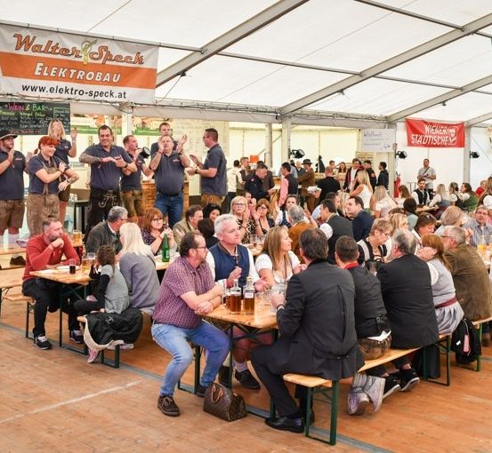 Eine große Gruppe von Menschen versammelt sich in einem Zelt, einige sitzen auf Bänken und andere stehen. Sie sind in traditioneller bayerischer Kleidung gekleidet und scheinen Getränke und Essen zu genießen. Das Zelt ist mit Bannern und Schildern geschmückt, und es gibt eine Bühne mit einem Mann, der auftritt.