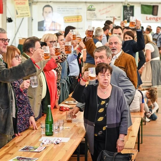 Eine Gruppe von Menschen bei einer Outdoor-Veranstaltung, die Gläser mit Bier zum Anstoßen heben. Einige sitzen an langen Tischen, andere stehen dahinter. Die Umgebung scheint ein Festival zu sein, mit Bannern und einem großen Porträt im Hintergrund.