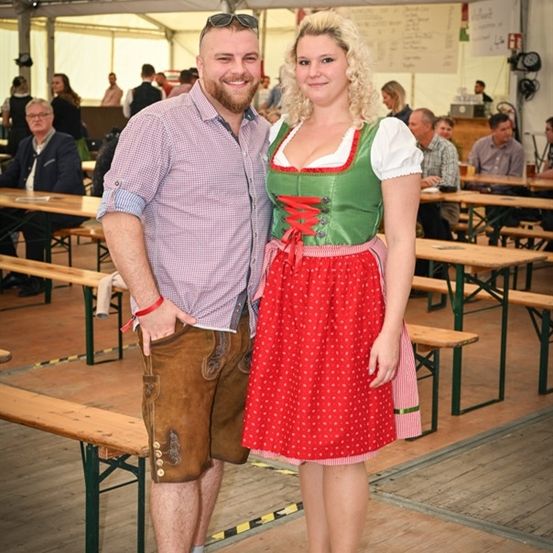 Ein Mann und eine Frau in traditioneller deutscher Kleidung stehen in einem Bierzelt und lächeln für die Kamera. Hinter ihnen sind Tische und Bänke mit Menschen gefüllt.