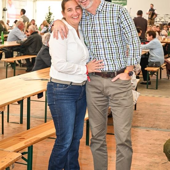 Ein lächelndes Paar steht in einem überfüllten Biergarten, mit hölzernen Bänken und Tischen im Hintergrund. Der Mann trägt ein kariertes Hemd, und die Frau trägt eine weiße Bluse.