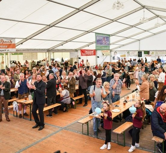 Eine große Gruppe von Menschen versammelt sich in einem Zelt für eine Veranstaltung, einige sitzen an Tischen mit Getränken und andere stehen und klatschen. Banner hängen von der Decke, und der Boden ist aus Holz.