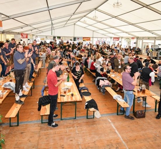 Eine große Menschenmenge versammelt sich in einem Zelt für eine Veranstaltung, sitzt an langen Tischen und trinkt Bier. Das Zelt ist mit Bannern dekoriert und hat einen Holzboden.