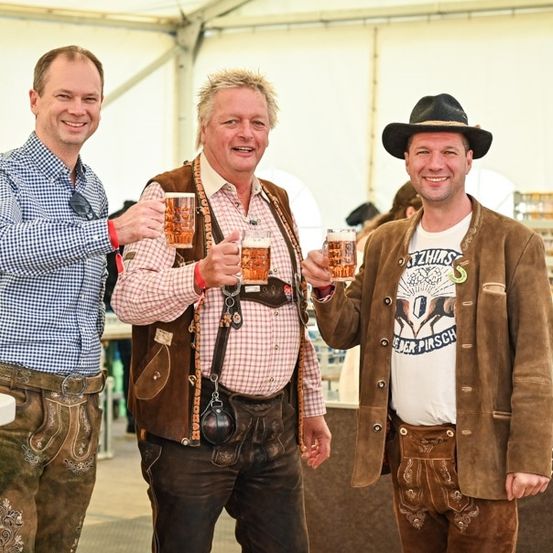 Drei Männer in traditioneller bayerischer Kleidung posieren für ein Foto mit Bierkrügen in der Hand. Der Mann links trägt ein kariertes Hemd und eine Hose mit Gürtel. Der Mann in der Mitte hat eine Weste und ein kariertes Hemd. Der Mann rechts trägt eine braune Jacke und einen Hut.