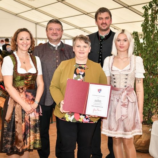 Eine Gruppe von Menschen in traditioneller bayerischer Kleidung steht zusammen, wobei eine Frau ein Zertifikat hält. Der Ort scheint eine formelle Veranstaltung oder Preisverleihung zu sein.